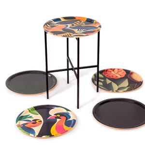 Może przedstawiać: Mały, okrągły stolik pomocniczy z czarną metalową ramą i blatem we wzory z pomarańczowymi, niebieskimi i czarnymi abstrakcyjnymi kształtami. Kilka dekoracyjnych tac z czarnymi powierzchniami i kolorowymi wzorami kwiatowymi i ptasimi jest ułożonych wokół stołu.