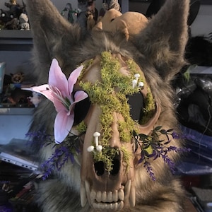 May include: A detailed close-up of a wolf-like mask. The mask features a skull design with faux fur, adorned with moss, small mushrooms, and purple and pink flowers. The mask has large, pointed ears and a detailed jawline.