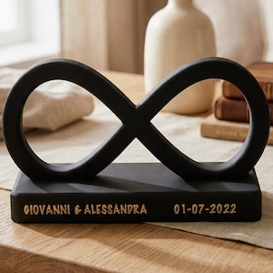 May include: A black infinity symbol sculpture on a rectangular base. The base has the names "GIOVANNI & ALESSANDRA" and the date "01-07-2022" in gold lettering. The sculpture is displayed on a wooden table, with a vase and books in the background.
