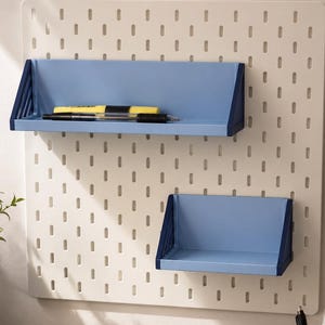 Op de afbeelding: Een witte pegboard met twee blauwe planken. De planken zijn gemonteerd op de pegboard, die een raster van kleine gaten heeft. De bovenste plank bevat een gele markeerstift en een zwarte pen. Een kleine plant in een witte pot en een notitieboek liggen op het bureau.