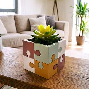 Può includere: Un vaso quadrato con un design a pezzi di puzzle, caratterizzato da pannelli opachi nei toni del terracotta, verde salvia, bianco e giallo senape. Una pianta succulenta verde brillante con punte gialle è piantata nel contenitore cubico.