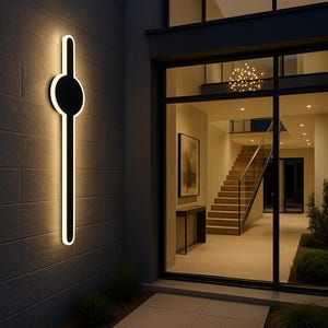 May include: Exterior wall with a modern black and white light fixture. The fixture has a long, vertical design with a circular element at the top. The light is illuminated, creating a warm glow against the dark wall.