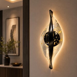 May include: A modern wall sconce with a clear, leaf-shaped glass design, accented with gold leaf and a black central element. The sconce has a black, curved support and emits a warm, ambient light, creating a decorative and functional lighting fixture.