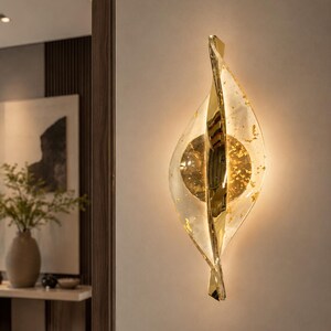May include: A modern wall sconce with a gold and clear glass design. The sconce has a leaf-like shape with a central gold disc and gold leaf accents. It is mounted on a neutral-colored wall and provides ambient lighting.