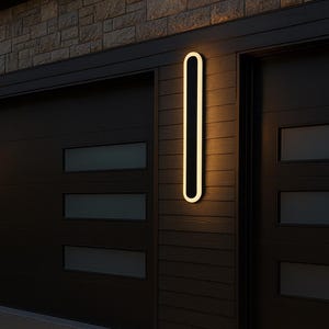 May include: Exterior wall-mounted light fixture with a vertical, elongated oval design. The light has a black center and a bright, warm white border. It is mounted on a dark brown wooden wall next to a black garage door.