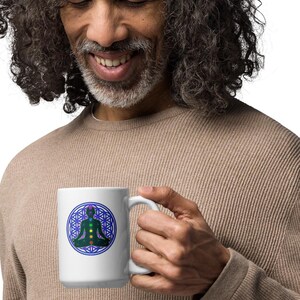 May include: White ceramic mug featuring a blue and green geometric design with a meditating figure. The mug is held by a person wearing a tan long-sleeve shirt. The design is centered on the mug.