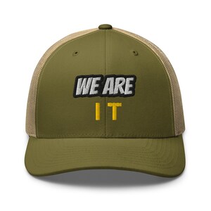 May include: Olive green trucker hat with the embroidered text "WE ARE IT" in white and black, and the word "IT" in yellow. The hat has a mesh back and a curved brim.