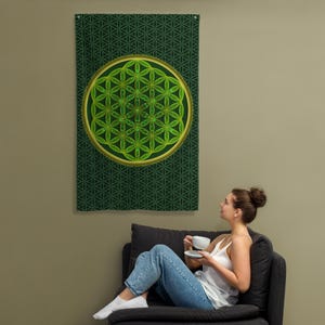 Könnte beinhalten: Ein grünes Wandbehang mit geometrischem Muster und einem goldumrandeten Kreis mit einem grünen Blumendesign. Der Wandteppich hängt an einer Wand. Eine Frau sitzt auf einem schwarzen Sofa und betrachtet den Wandteppich.
