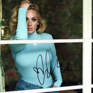 Puede incluir: Retrato de una mujer con cabello rubio, vestida con un suéter de cuello alto azul claro y jeans azules, apoyada en una ventana. La imagen incluye un autógrafo en tinta negra. El fondo está borroso.