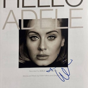 Op de afbeelding: Een bladmuziekboek getiteld HELLO ADELE voor makkelijke piano, met een zwart-witportret van Adele in het midden. De omslag bevat een handtekening in blauwe inkt, tekstcredits voor het nummer en logo's van Universal, Hal Leonard en EMI.