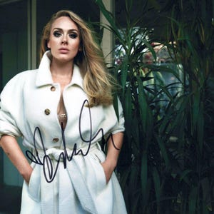May include: A glossy photograph of Adele wearing a long-sleeved, off-white coat with a wide collar and three brown buttons. The coat features a matching tie belt at the waist. A black ink autograph is written across the center of the image.