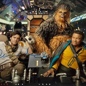 May include: A Star Wars film still showing three characters in a spaceship cockpit. Chewbacca, a Wookiee, is in the background. Oscar Isaac and a man in a yellow shirt are in the foreground. The image is signed in blue ink.