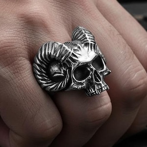 Może przedstawiać: Srebrny pierścień z czaszką z zakrzywionymi rogami i szczegółowymi rysami twarzy. Pierścień jest noszony na palcu, prezentując skomplikowany design i gotycki styl. Pierścień jest wyrazistym elementem.