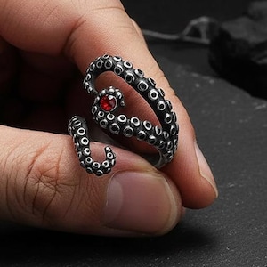 May include: A silver-colored ring in the shape of an octopus tentacle, with a small red gemstone at the center. The ring is detailed with suction cups and is shown on a person's finger.