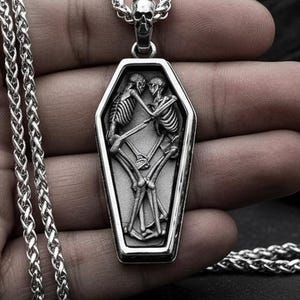 Op de afbeelding: Zilverkleurige hangerketting met een doodskistvormige hanger met een gegraveerd ontwerp van twee omarmende skeletten. De hanger hangt aan een fijne ketting. De ketting wordt gepresenteerd tegen een donkere achtergrond.