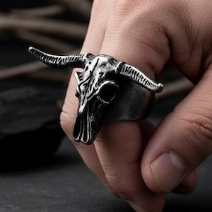 May include: A silver-colored ring featuring a bull skull design. The ring has detailed horns and skull features. The ring is worn on a finger, showcasing the intricate craftsmanship and gothic style of the jewelry.