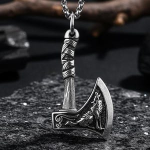 Puede incluir: Collar con colgante de hacha plateada. El colgante presenta una cabeza de hacha detallada con un diseño de lobo y árbol. El collar tiene una cadena con un cierre de mosquetón. El colgante mide aproximadamente 3,8 cm de largo.