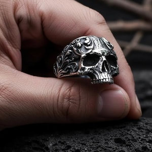 Op de afbeelding: Een zilveren schedelring met ingewikkelde bloemendetails. De ring heeft een gedetailleerd schedelontwerp met open oogkassen en een volledige set tanden. De ring wordt tegen een donkere achtergrond gehouden.