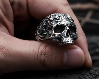 Könnte beinhalten: Ein silberner Totenkopfring mit aufwendigen floralen Details. Der Ring zeigt ein detailliertes Totenkopfdesign mit offenen Augenhöhlen und einem vollständigen Gebiss. Der Ring wird vor einem dunklen Hintergrund gehalten.