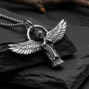 May include: A silver-colored angel pendant necklace. The pendant features an angel with outstretched wings. The necklace is on a dark, textured surface, with a small branch in the background. The chain is a linked style.