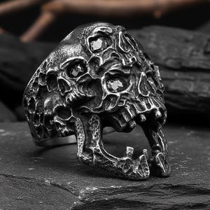 May include: A detailed, silver-toned skull ring with multiple small skulls forming the main design. The ring has a textured, weathered appearance, with dark accents in the crevices. The open mouth of the skull is a prominent feature.