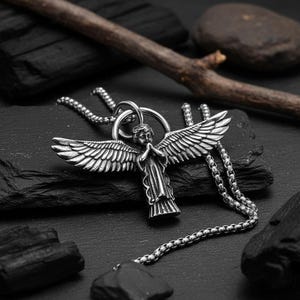 May include: A silver-colored angel pendant necklace. The pendant features an angel with outstretched wings and clasped hands. The necklace has a chain with a box link design. The angel pendant is displayed on a black, textured surface.