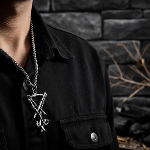 Puede incluir: Collar con colgante plateado con un dise&ntilde;o geom&eacute;trico &uacute;nico. El colgante cuelga de una cadena trenzada y se lleva sobre una camisa negra. El fondo presenta una pared de ladrillos oscuros y algunas ramas.