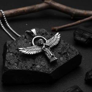May include: A silver-colored angel pendant necklace. The pendant features an angel with outstretched wings. The necklace is on a dark, textured surface. The angel has detailed wings and a halo.
