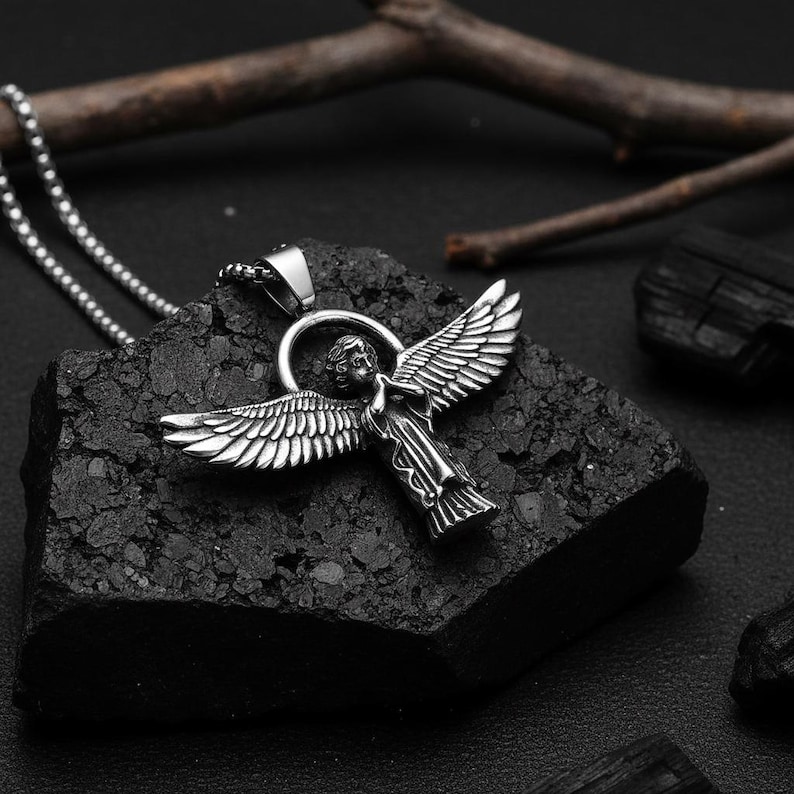May include: A silver-colored angel pendant necklace. The pendant features an angel with outstretched wings. The necklace is on a dark, textured surface. The angel has detailed wings and a halo.