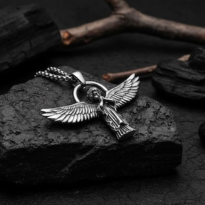 May include: A silver-colored angel pendant with outstretched wings, suspended from a ball chain necklace. The angel figure is detailed, with visible feathers and clothing. The pendant rests on a dark, textured surface.
