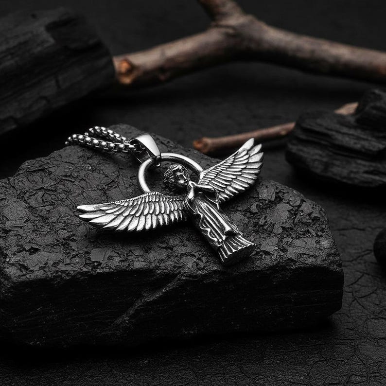 May include: A silver-colored angel pendant with outstretched wings, suspended from a ball chain necklace. The angel figure is detailed, with visible feathers and clothing. The pendant rests on a dark, textured surface.