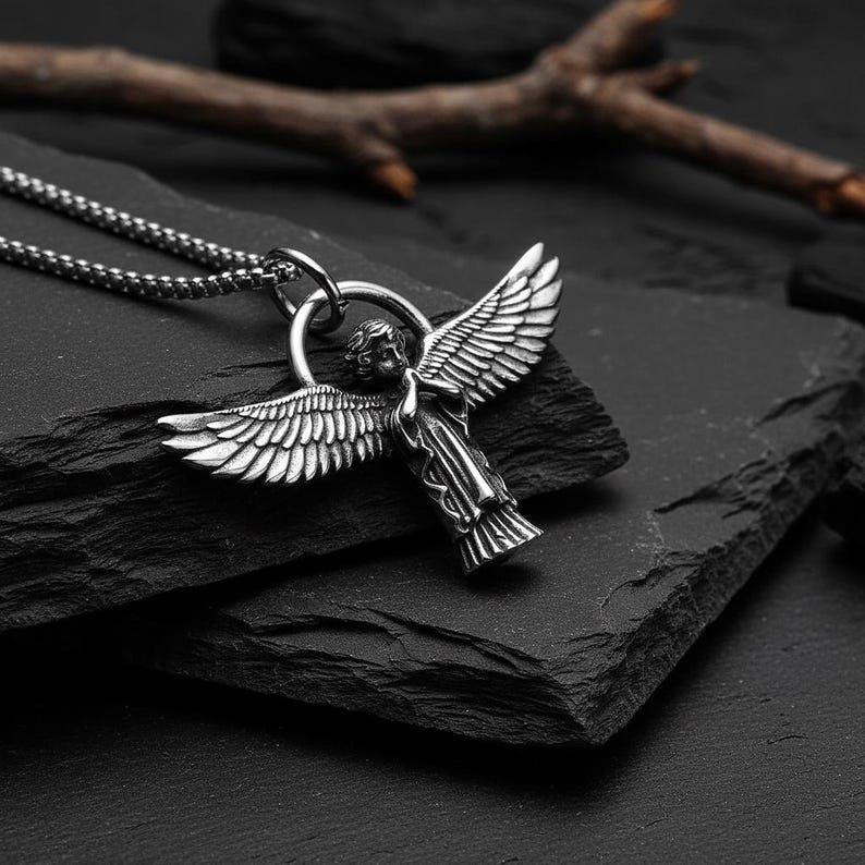 May include: A silver-colored angel pendant necklace. The pendant features an angel with outstretched wings. The necklace is on a dark, textured surface, with a small branch in the background. The chain is a linked style.
