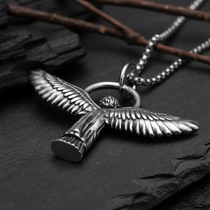 May include: A silver-colored pendant necklace featuring a detailed angel with outstretched wings. The pendant is suspended from a beaded chain. The necklace is set against a dark, textured background with twigs.