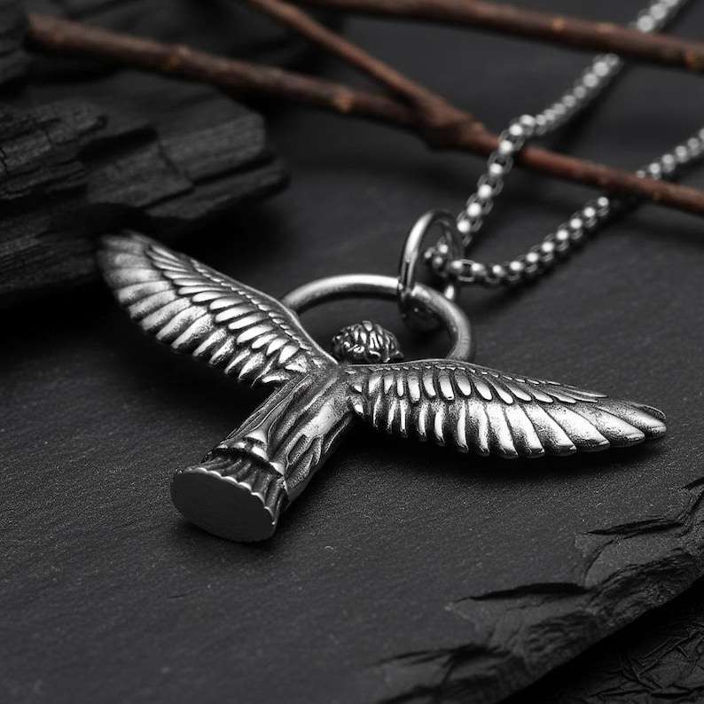 May include: A silver-colored pendant necklace featuring a detailed angel with outstretched wings. The pendant is suspended from a beaded chain. The necklace is set against a dark, textured background with twigs.