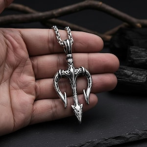 May include: Silver-toned trident pendant necklace. The pendant features a three-pronged design with intricate detailing. The necklace has a chain with a textured, linked pattern. The pendant is held in a hand against a dark background.