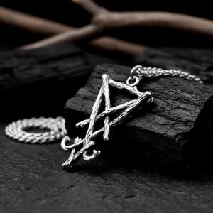 Puede incluir: Collar con colgante plateado con un dise&ntilde;o abstracto en forma de rama. El colgante cuelga de una fina cadena. El collar se muestra sobre una superficie oscura y texturizada. El metal plateado contrasta con el fondo oscuro.