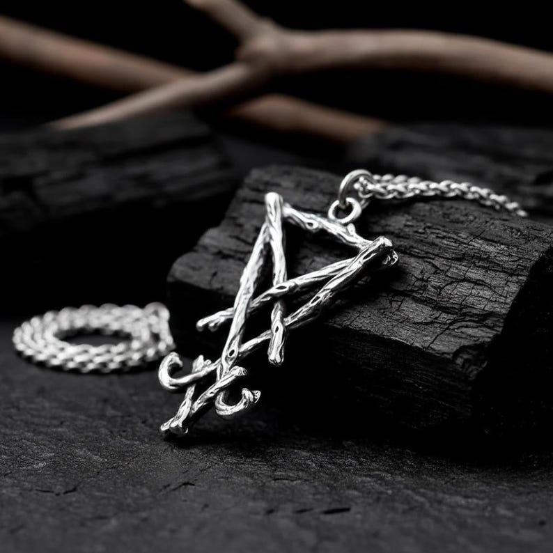 Puede incluir: Collar con colgante plateado con un dise&ntilde;o abstracto en forma de rama. El colgante cuelga de una fina cadena. El collar se muestra sobre una superficie oscura y texturizada. El metal plateado contrasta con el fondo oscuro.