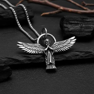 May include: A silver-colored angel pendant necklace. The pendant features an angel with outstretched wings and clasped hands. The necklace has a chain with a ball-like design. The background is dark, with a few pieces of black rock.