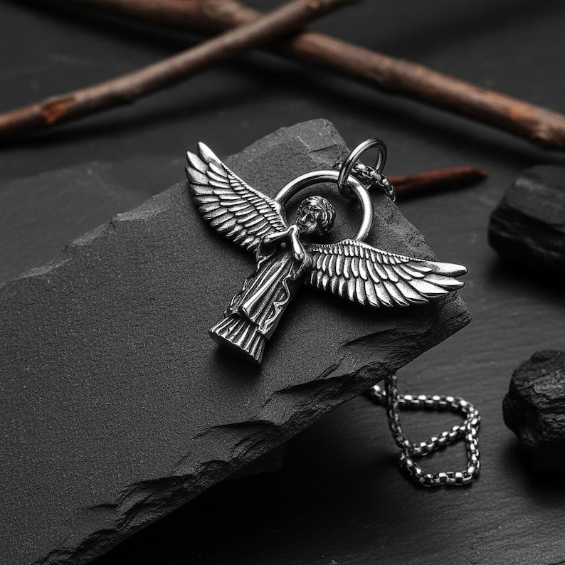 May include: A silver-colored angel pendant necklace. The pendant features an angel with outstretched wings, attached to a chain. The necklace is displayed on a dark gray, textured surface, with a few dark rocks and twigs in the background.