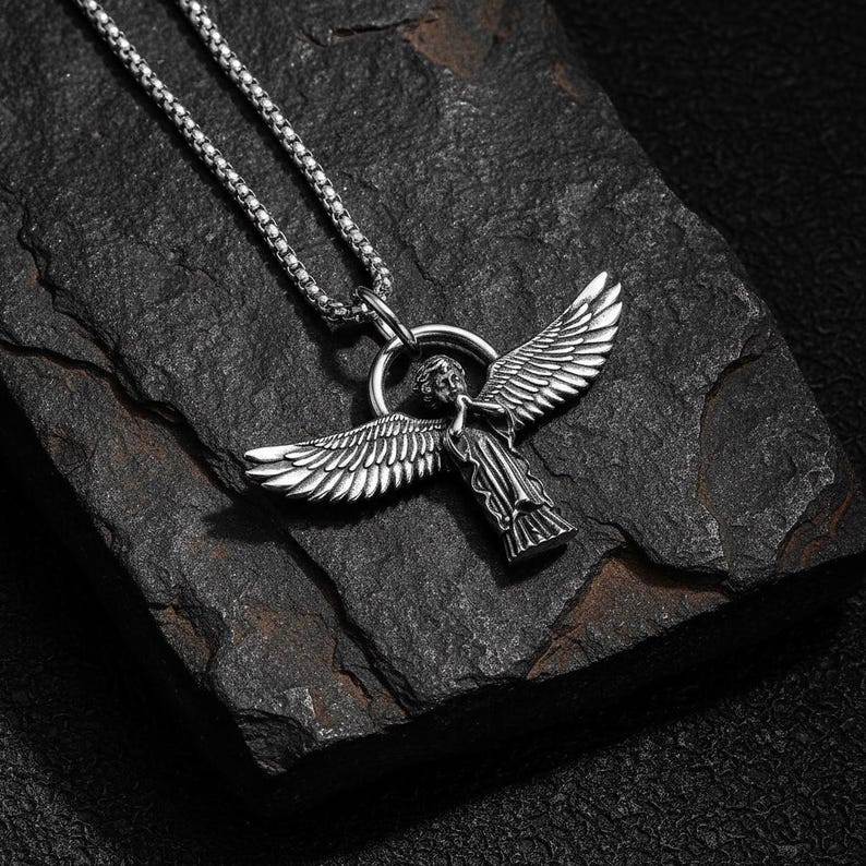 May include: Silver-toned angel pendant necklace. The pendant features an angel with outstretched wings, suspended from a chain. The necklace is displayed on a dark, textured surface, creating a striking contrast.