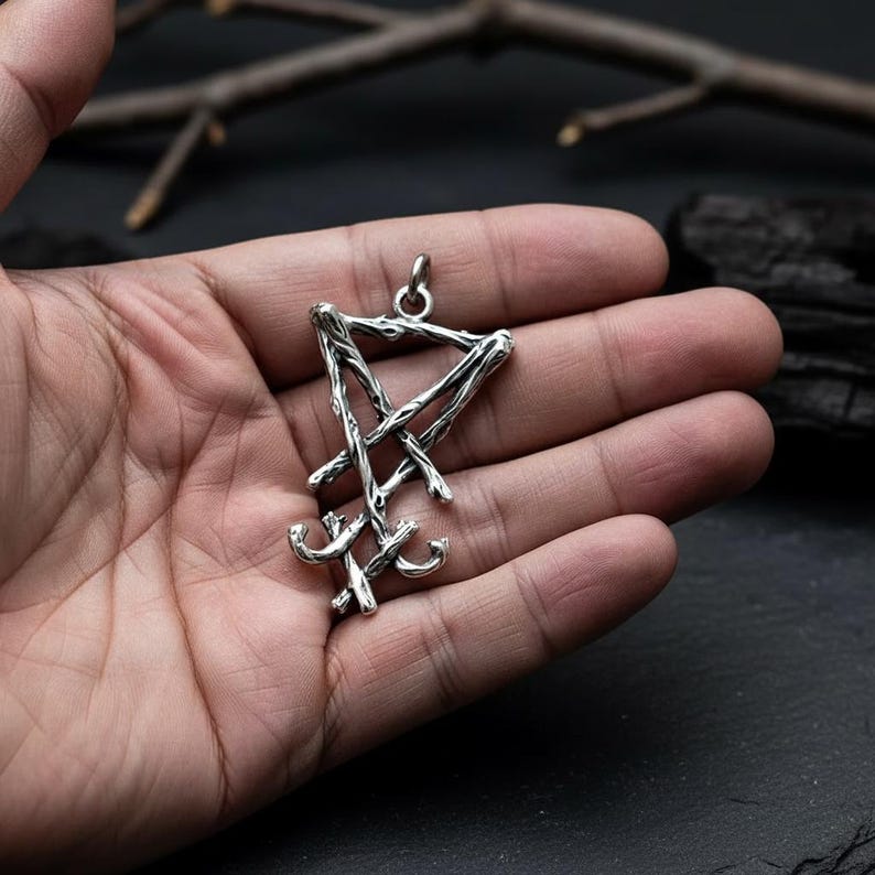 Puede incluir: Colgante plateado con el dise&ntilde;o del Sello de Lucifer. El colgante tiene una apariencia texturizada y r&uacute;stica, con un pentagrama estilizado y un s&iacute;mbolo de cruz invertida. El colgante se sostiene en una mano, mostrando sus intrincados detalles.