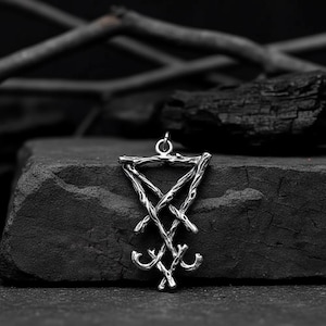 Puede incluir: Un colgante plateado con el sigilo de Lucifer, un s&iacute;mbolo del ocultismo. El colgante est&aacute; hecho de l&iacute;neas entrelazadas y cuelga de un peque&ntilde;o lazo. El fondo es una superficie oscura y texturizada con carb&oacute;n vegetal y ramas.