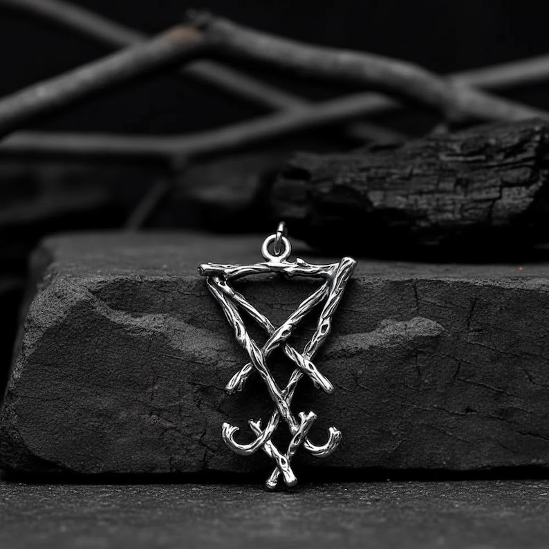 Puede incluir: Un colgante plateado con el sigilo de Lucifer, un s&iacute;mbolo del ocultismo. El colgante est&aacute; hecho de l&iacute;neas entrelazadas y cuelga de un peque&ntilde;o lazo. El fondo es una superficie oscura y texturizada con carb&oacute;n vegetal y ramas.