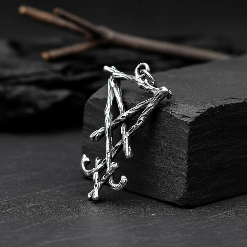 Puede incluir: Un colgante plateado con un dise&ntilde;o &uacute;nico en forma de ramita. El colgante de metal tiene un lazo para una cadena. El dise&ntilde;o es detallado, con una est&eacute;tica r&uacute;stica y natural. El colgante se muestra sobre una superficie oscura y texturizada.