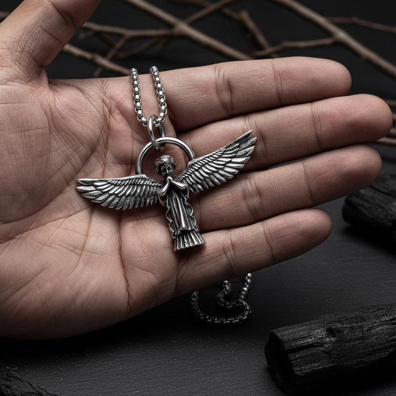 May include: A silver-colored angel pendant necklace. The pendant features an angel with outstretched wings and clasped hands, suspended from a chain. The necklace is displayed against a dark background.