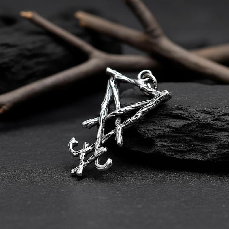 Puede incluir: Colgante plateado con forma de la letra "A". El colgante tiene un dise&ntilde;o texturizado, similar a una ramita. El fondo es oscuro, con una piedra negra y ramas de madera.