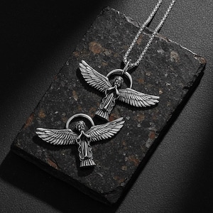 May include: Two silver-colored angel pendant necklaces. Each pendant features an angel with outstretched wings and a halo. The necklaces are displayed on a dark, textured surface, with one necklace chain visible.