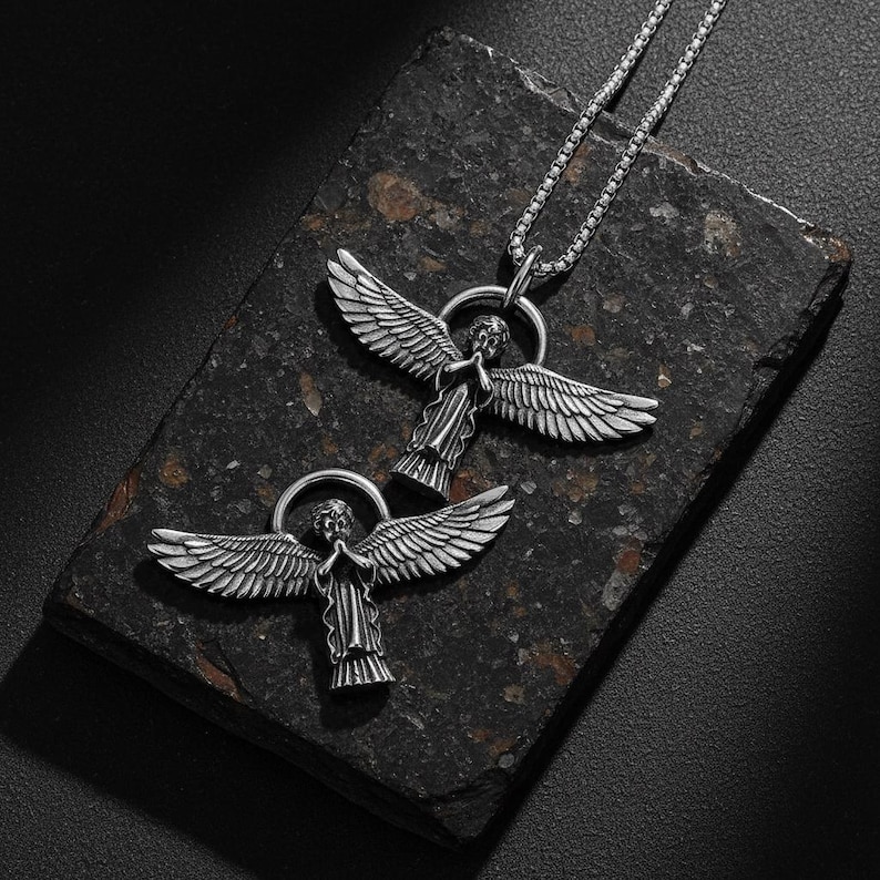 May include: Two silver-colored angel pendant necklaces. Each pendant features an angel with outstretched wings and a halo. The necklaces are displayed on a dark, textured surface, with one necklace chain visible.