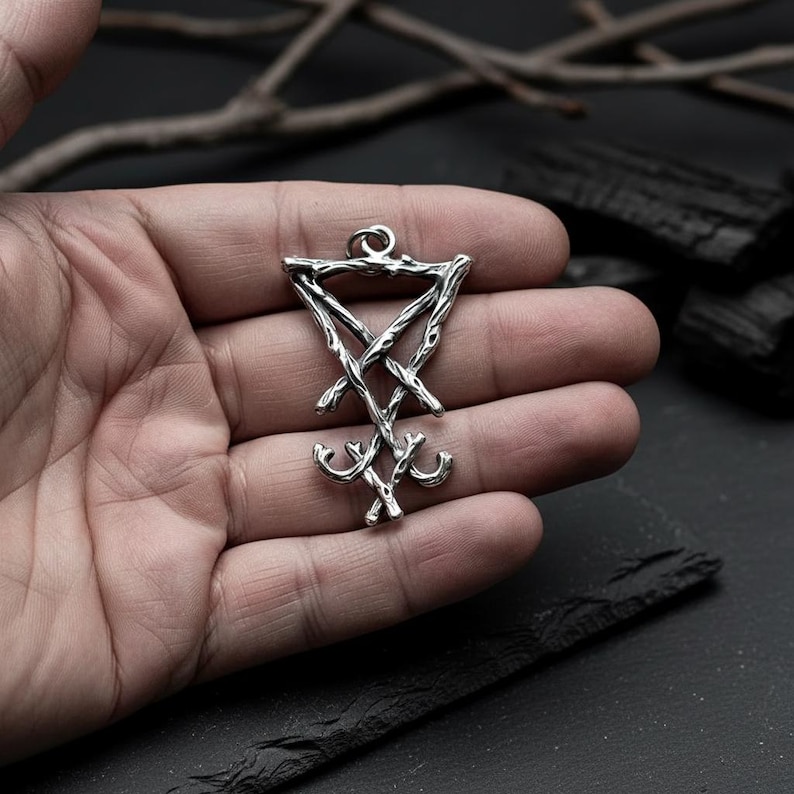 Puede incluir: Colgante plateado con el Sello de Lucifer, un s&iacute;mbolo de rebeli&oacute;n e iluminaci&oacute;n. El colgante tiene una parte superior triangular con un dise&ntilde;o de cruz y cruz invertida, y una parte inferior estilizada. El colgante se sostiene en una mano sobre un fondo oscuro.