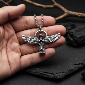 May include: Silver-toned angel pendant necklace. The pendant features an angel with outstretched wings and praying hands. The necklace has a chain with a circular bail. The angel pendant is held in a hand.
